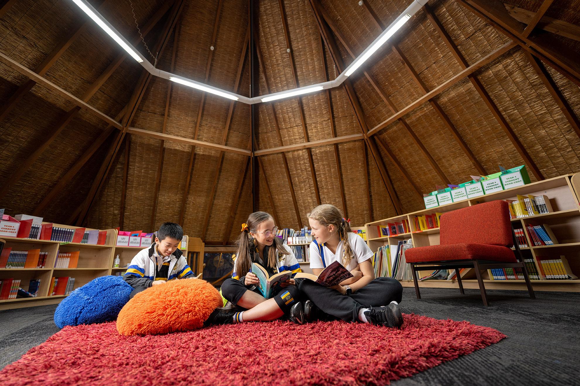 Three children sit on a red rug in a circular, warmly-lit room. They are engaged in reading books, surrounded by colorful bean bags and shelves filled with books.